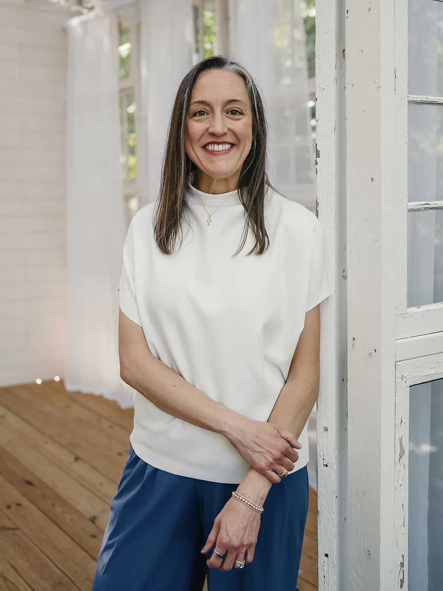 Smiling woman with brown hair wearing a white top and blue pants standing indoors by a white pillar