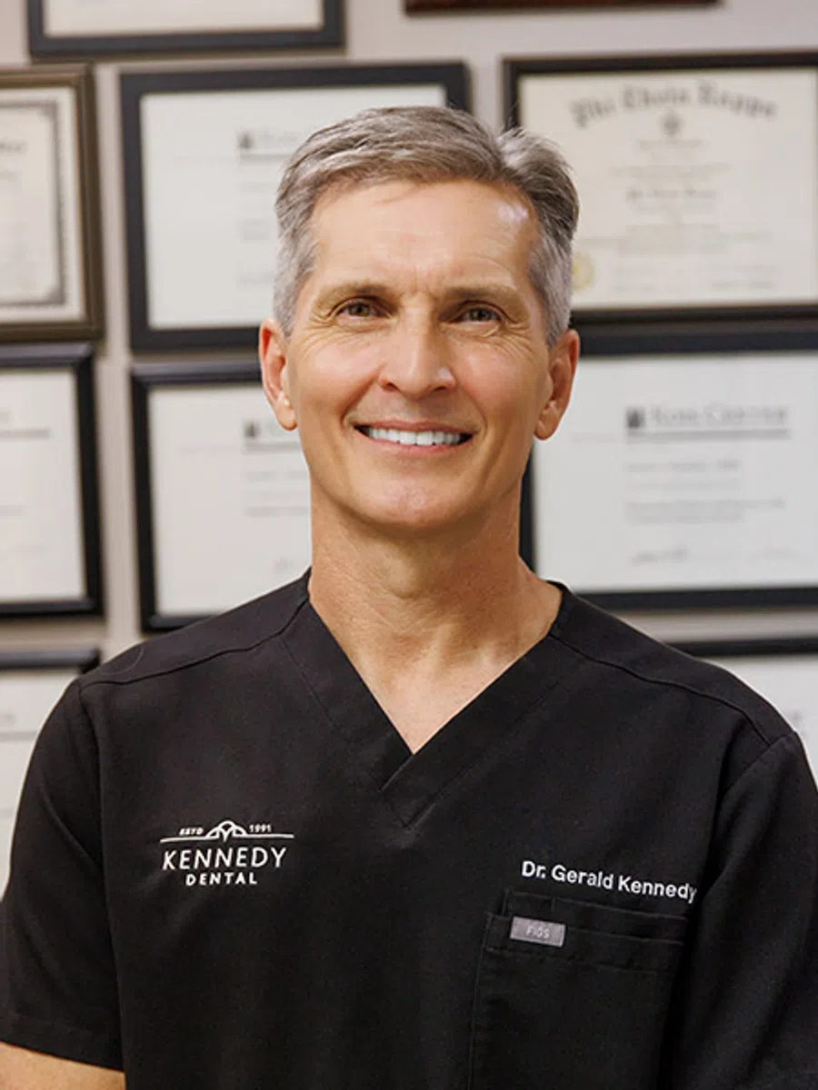 Smiling male dentist wearing black scrubs with Kennedy Dental logo and Dr. Kennedy name outside a building.