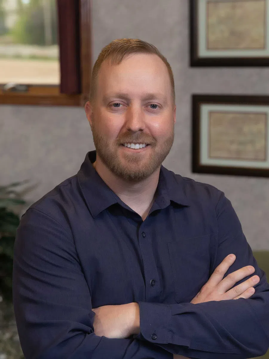 Smiling man with light beard wearing a navy blue shirt with arms crossed indoors by framed certificates.