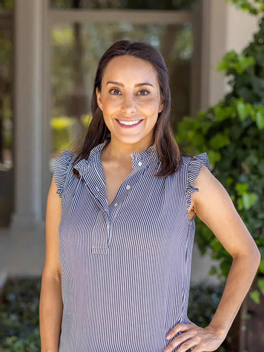 Smiling woman in striped sleeveless blouse posing outdoors with hand on hip and greenery in background