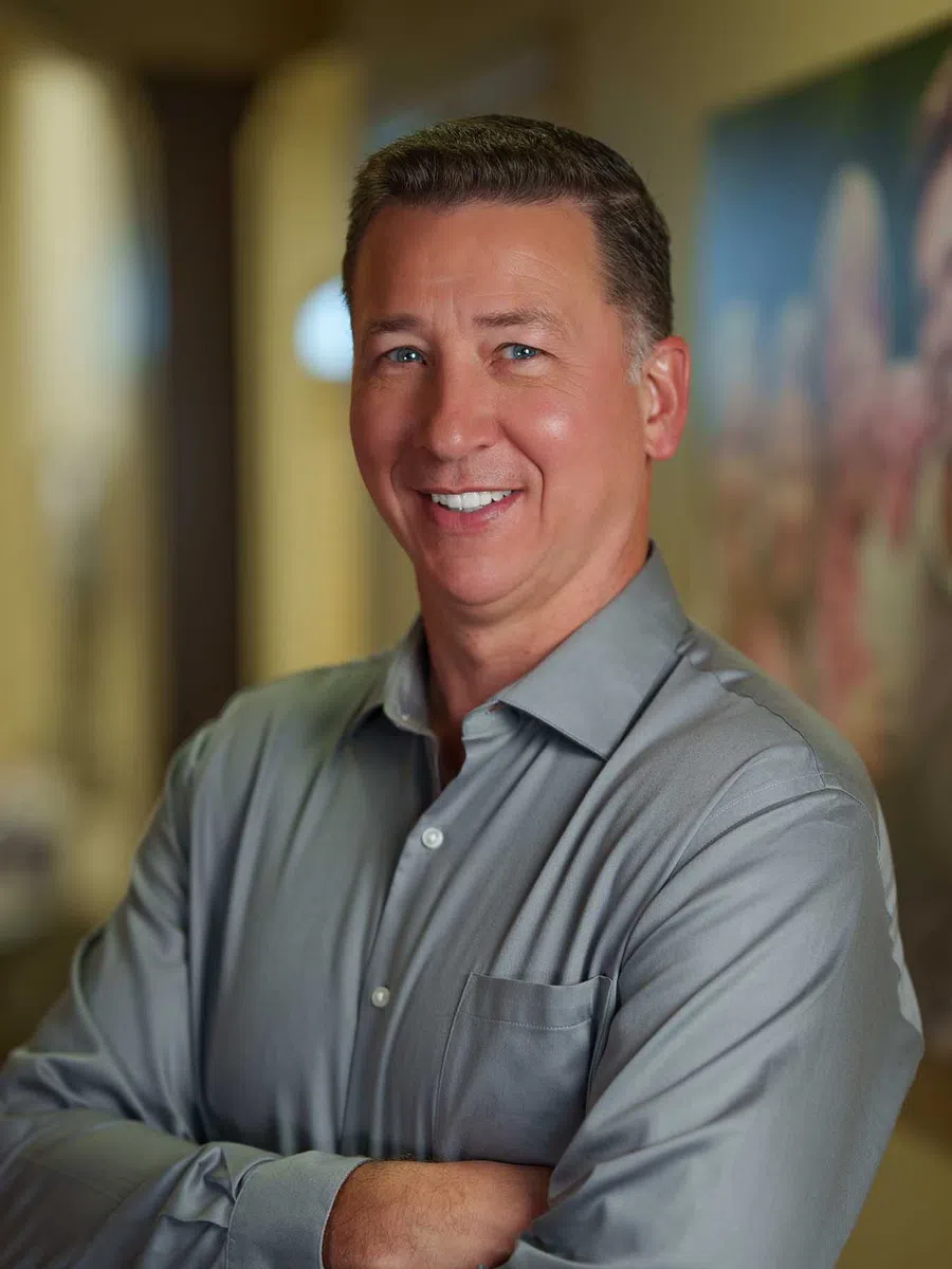 Smiling middle-aged man in a gray shirt with arms crossed standing indoors with blurred background.