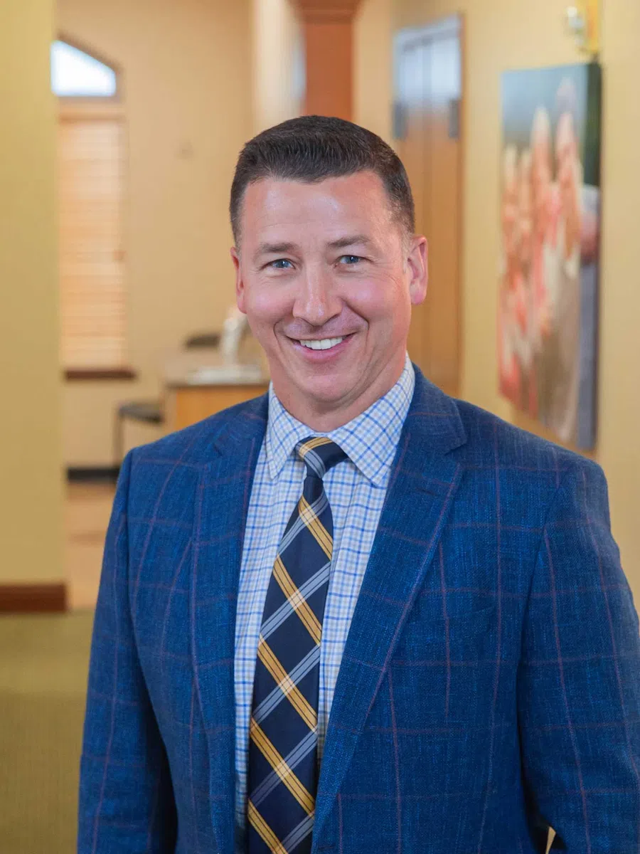 Smiling man in a blue checkered blazer and striped tie standing indoors with blurred background.