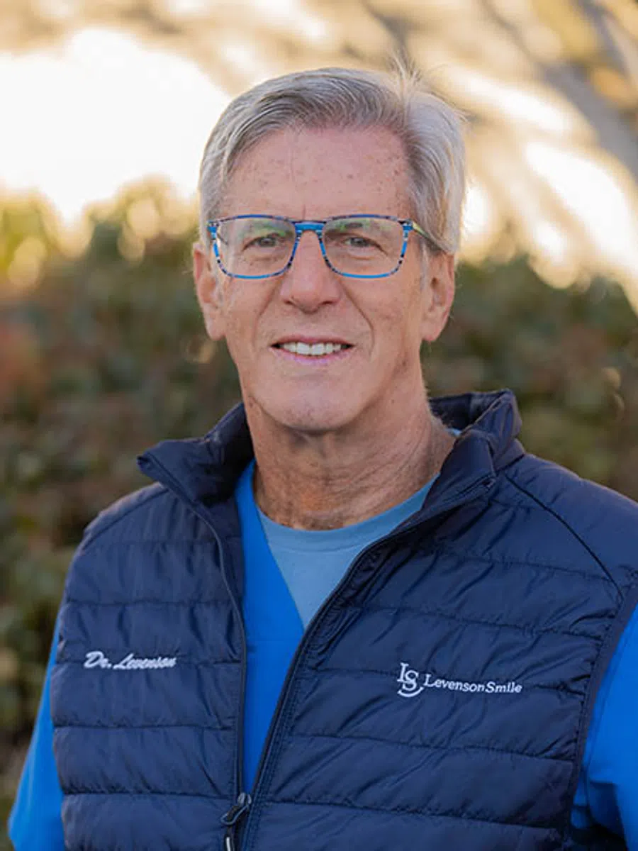 Older man wearing blue glasses and a navy Levenson Smile vest standing outdoors with a blurred background.