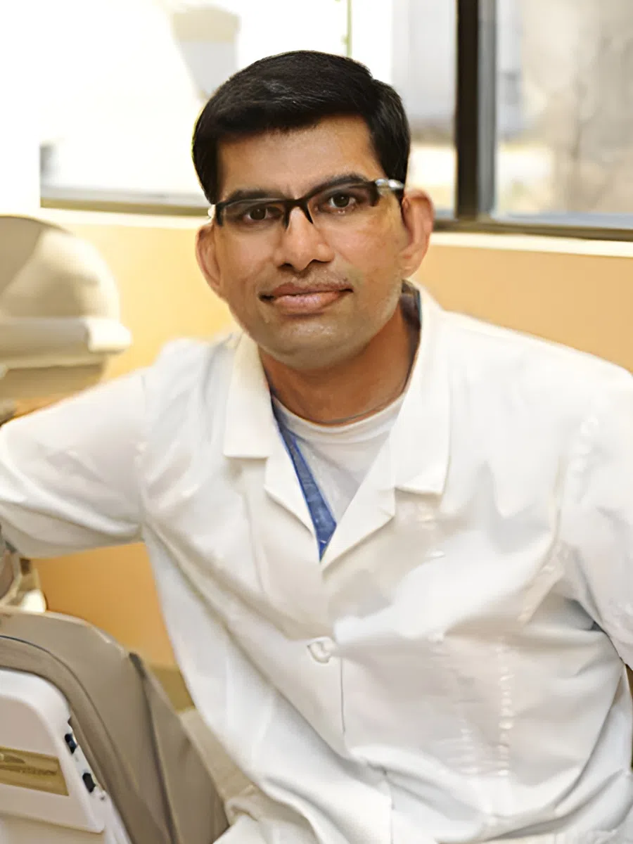 Male dentist wearing glasses and white coat leaning on dental chair with a window in the background