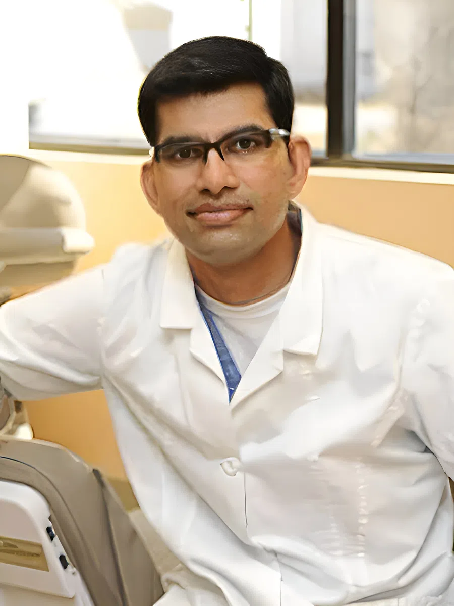 Male dentist wearing glasses and white coat in dental office sitting beside dental chair.