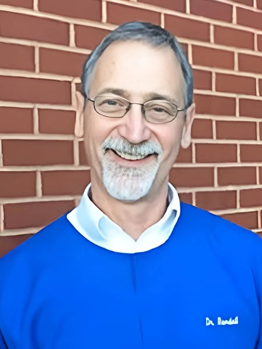 Senior man with glasses and gray beard wearing blue shirt with white collar smiling in front of brick wall.