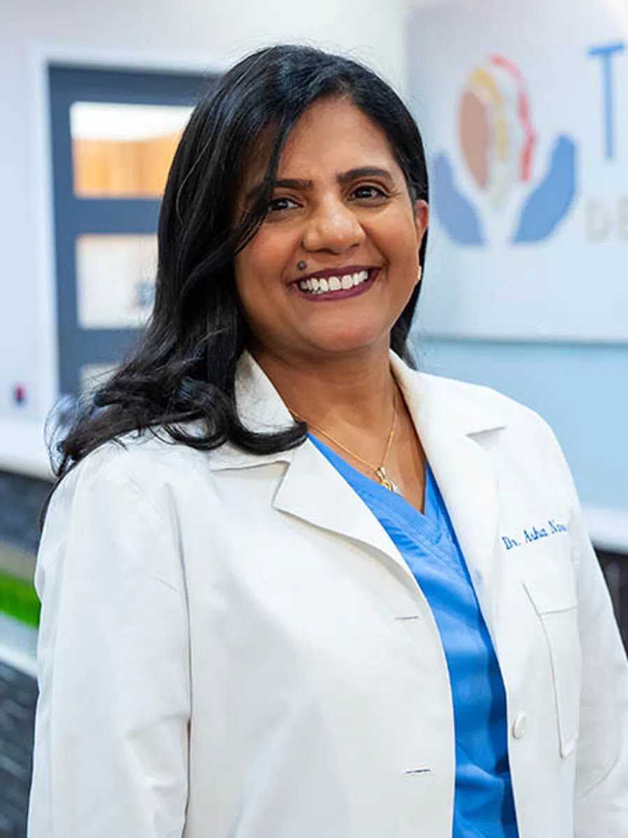 Smiling female doctor with long black hair in white coat and blue scrubs standing in medical office.