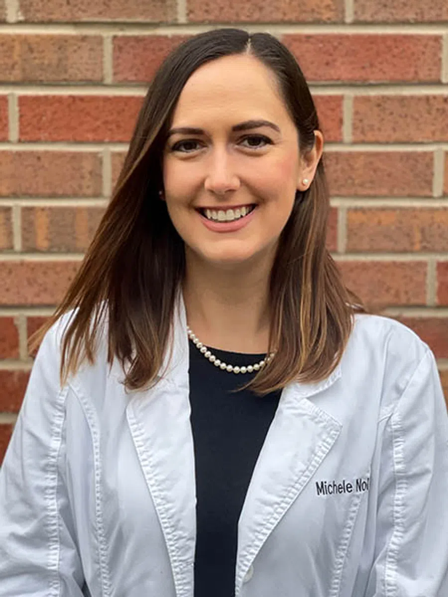 Smiling woman wearing a white lab coat, pearl necklace, black top, standing in front of a brick wall