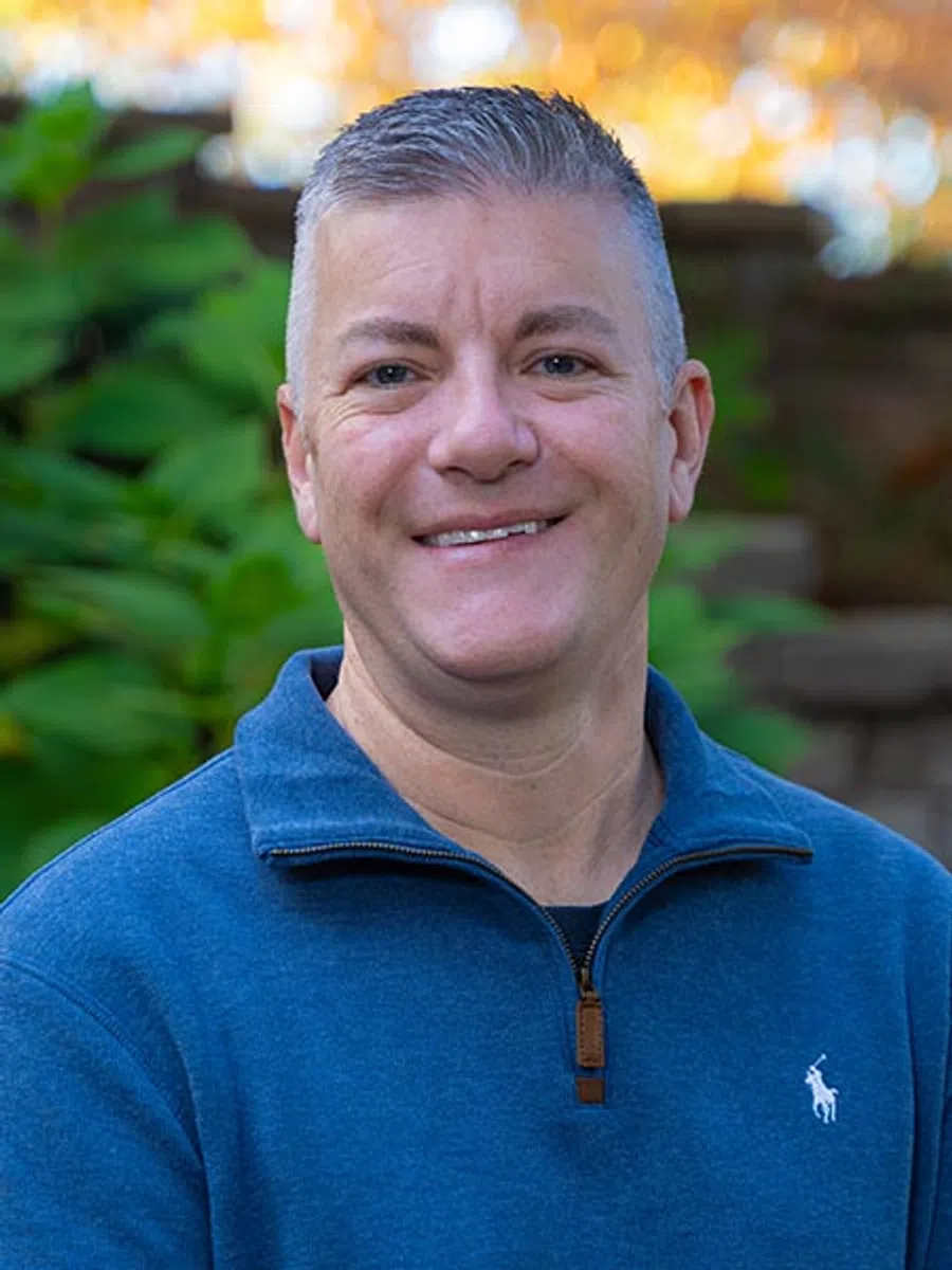 Smiling man wearing a blue quarter-zip sweater standing outdoors with green foliage and warm sunlight background.