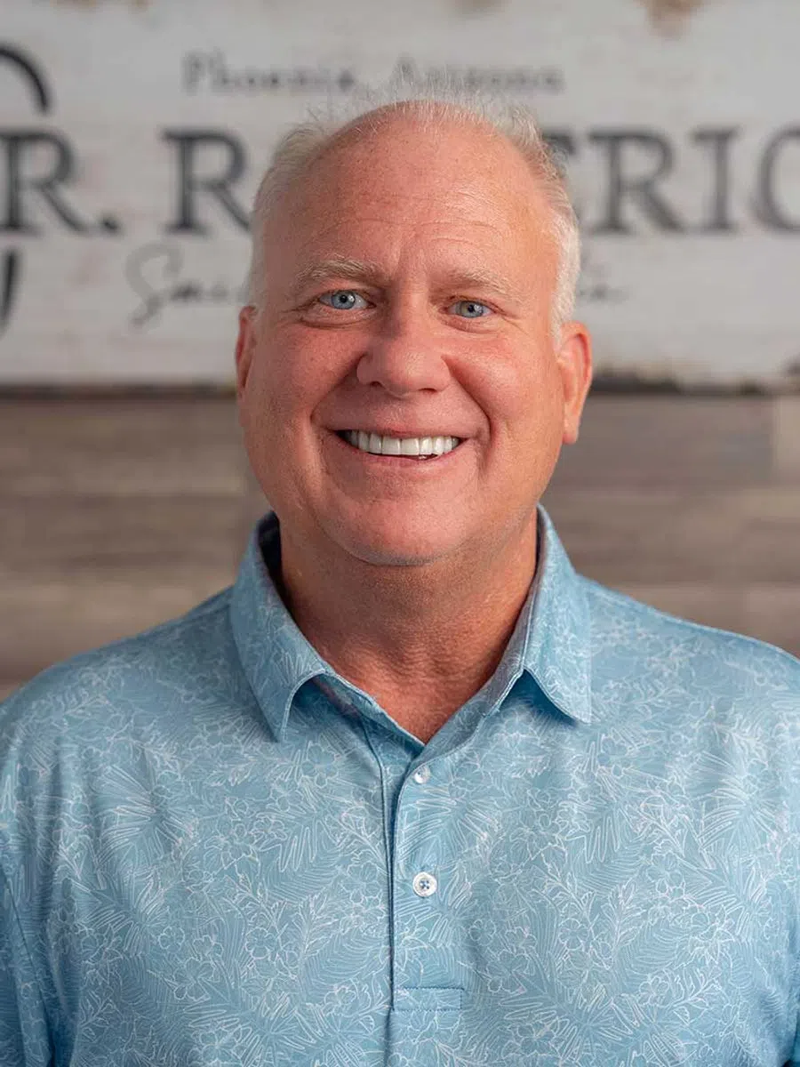 Smiling middle-aged man with light hair wearing a white jacket and blue patterned shirt in front of leafy background