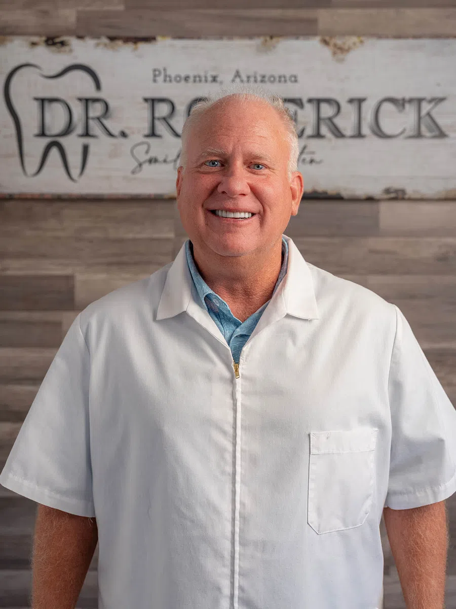 Smiling male dentist in white coat standing in front of a wooden sign reading Dr. Roderick in Phoenix Arizona.
