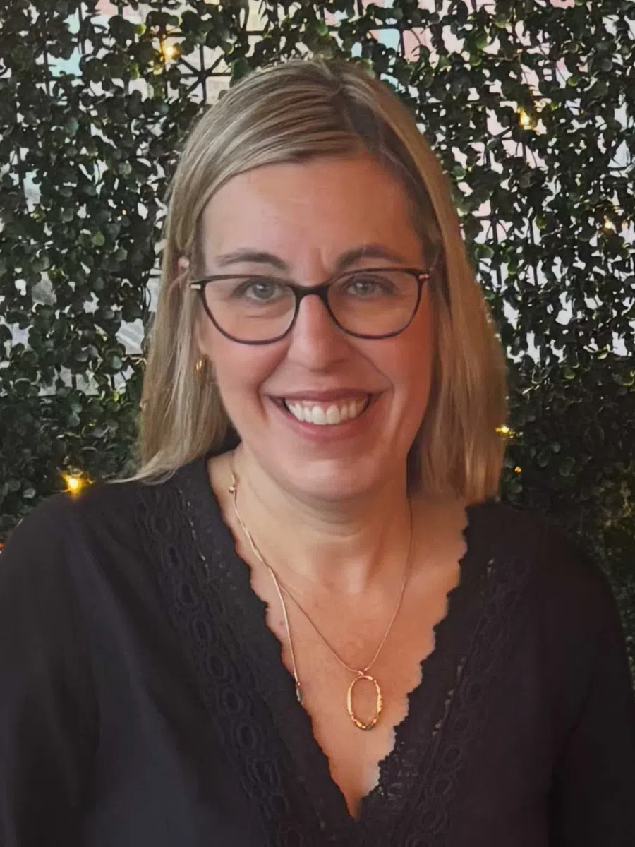 Smiling woman with blonde hair and glasses wearing a black lace top and gold necklace in front of leafy background.