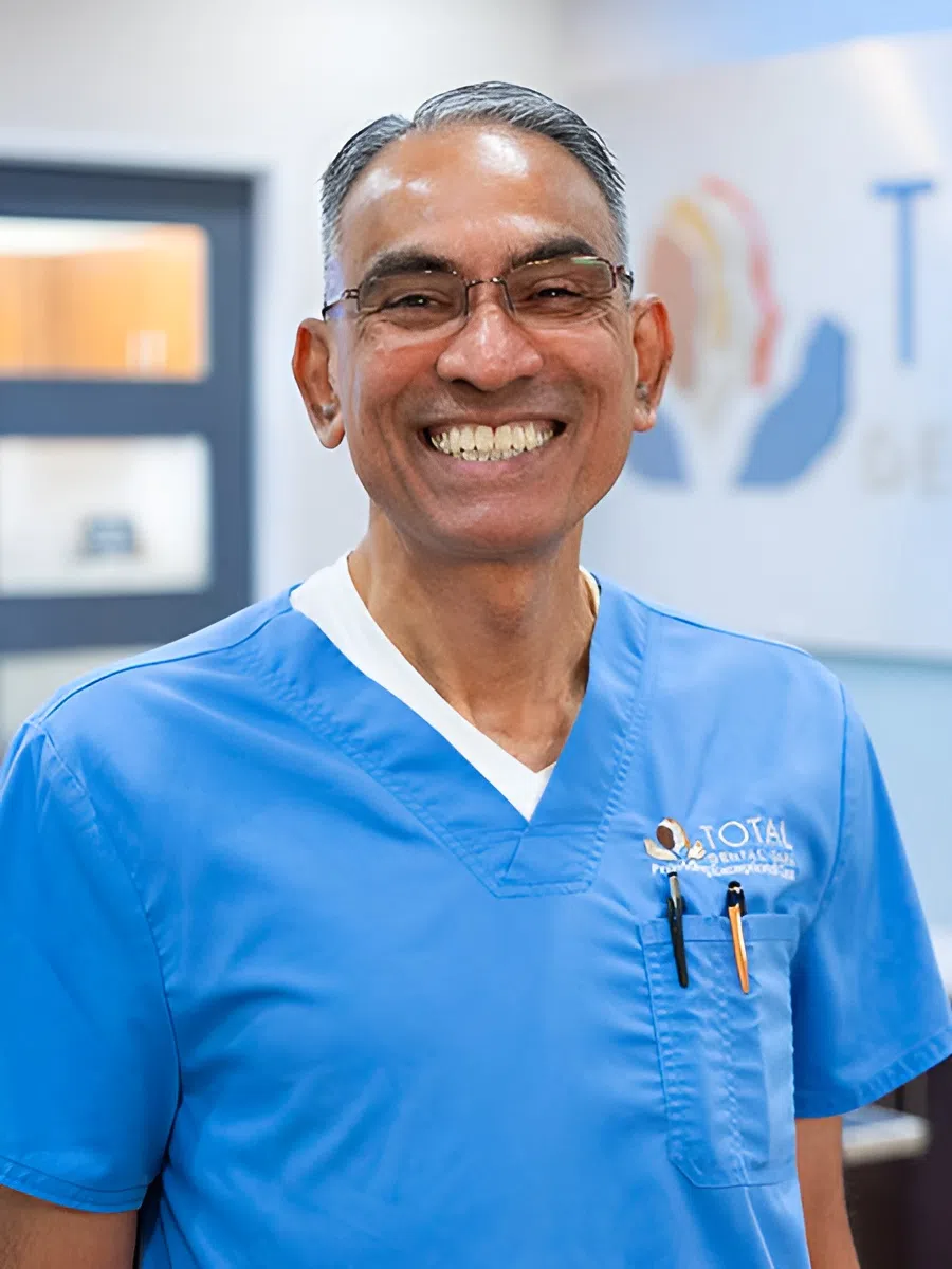 Smiling male dental professional wearing blue scrubs and glasses in a bright dental office.