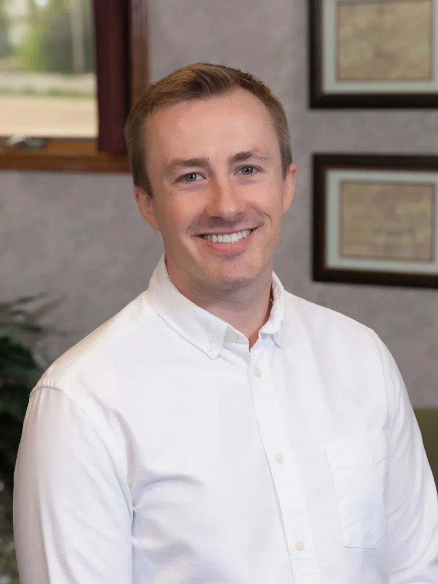 Smiling man in a white button-down shirt posing indoors with framed certificates on the wall behind him.