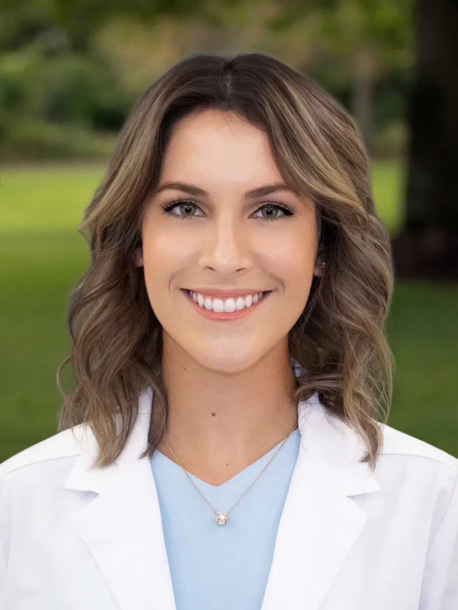 Smiling woman with shoulder-length wavy hair wearing a white lab coat and light blue top outdoors.