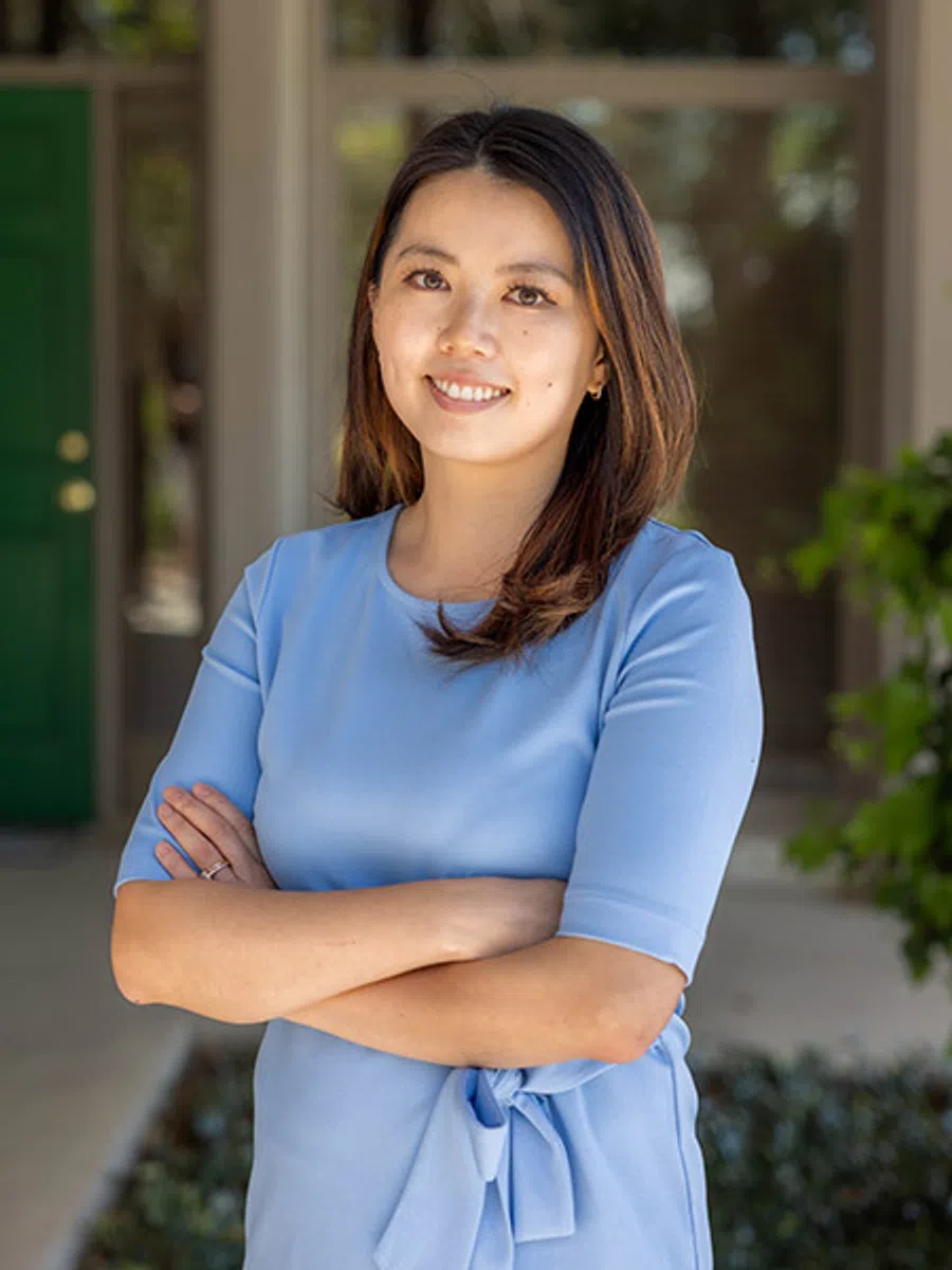 Smiling woman in light blue dress with crossed arms standing outdoors near green door and plants.