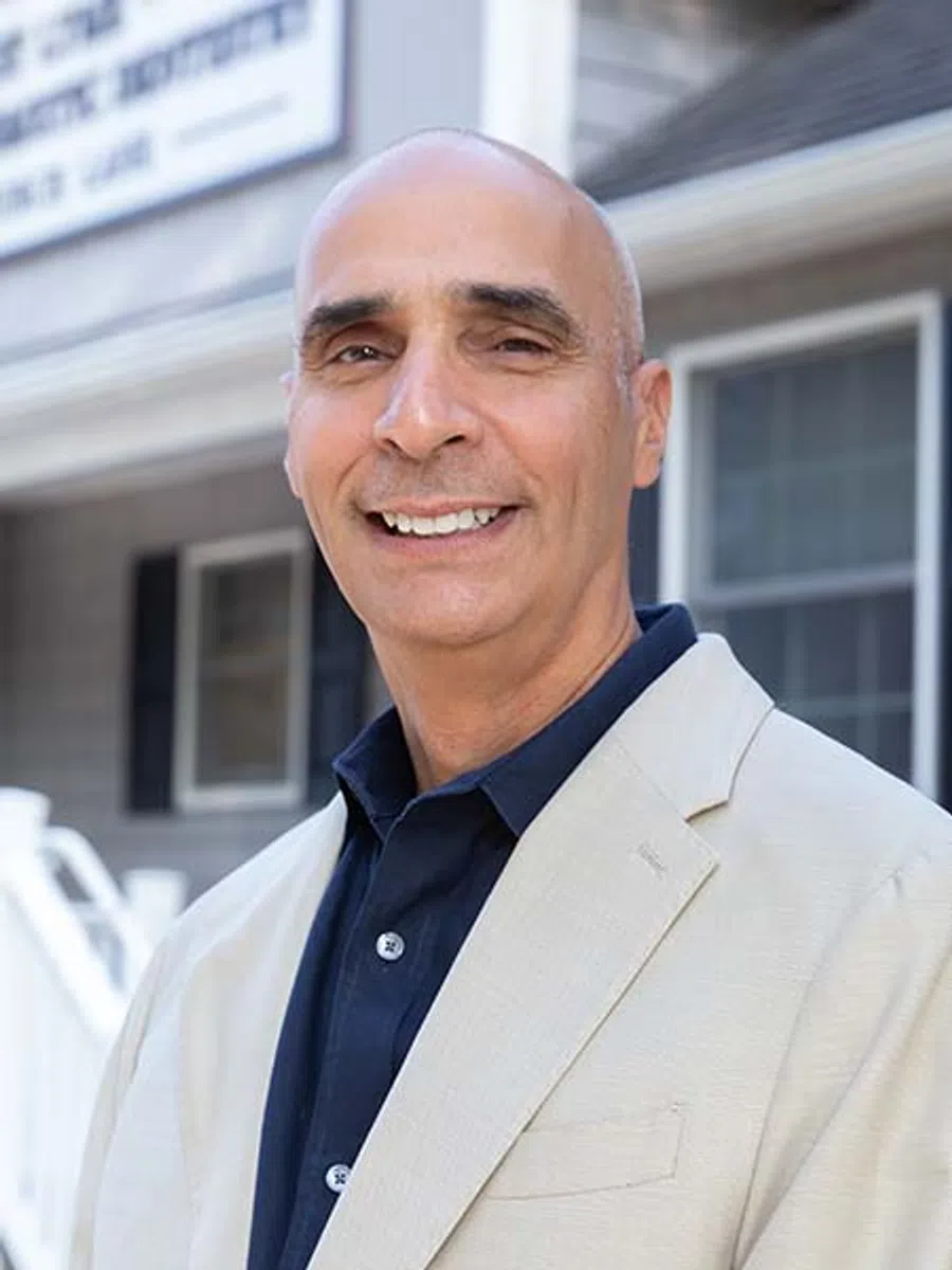 Smiling bald man in beige blazer and dark shirt standing outdoors in front of a house with windows and siding.