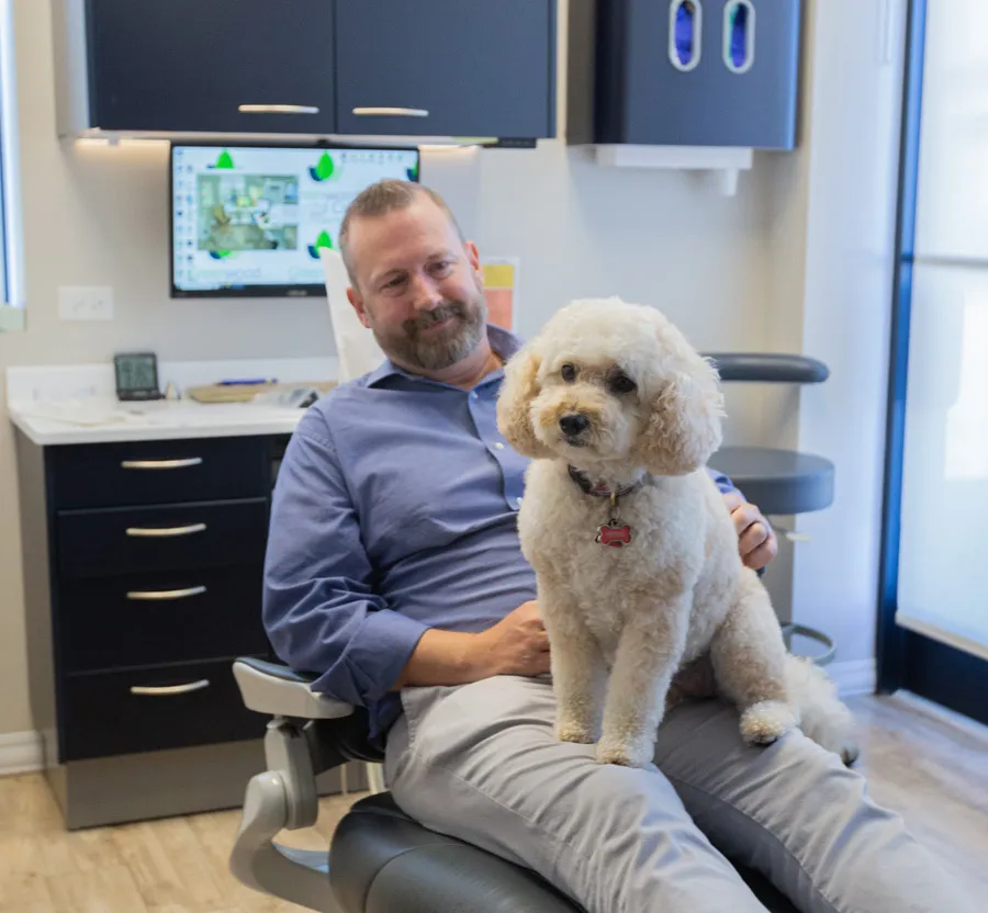 Man sitting in a dental chair with a small fluffy dog on his lap in a modern dental office.