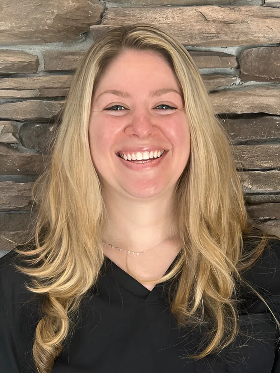 Smiling blonde woman with long hair wearing a black top against a brown stone wall background.