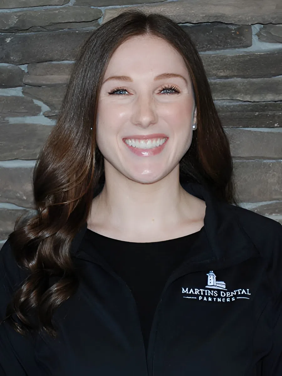 Smiling woman with long brown hair wearing a black Martins Dental Partners jacket against a stone wall.
