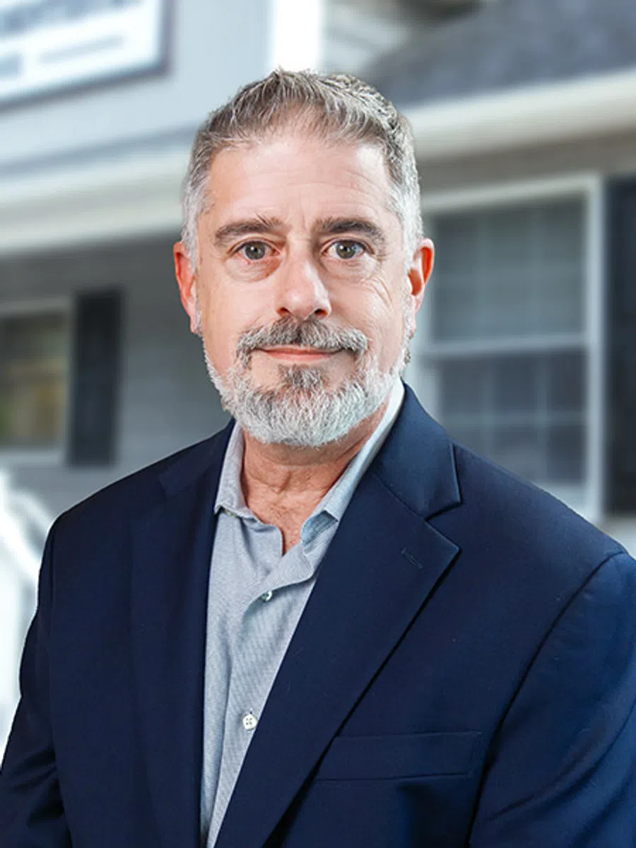 Mature man with gray beard wearing navy blazer and light gray shirt standing in front of a home exterior.