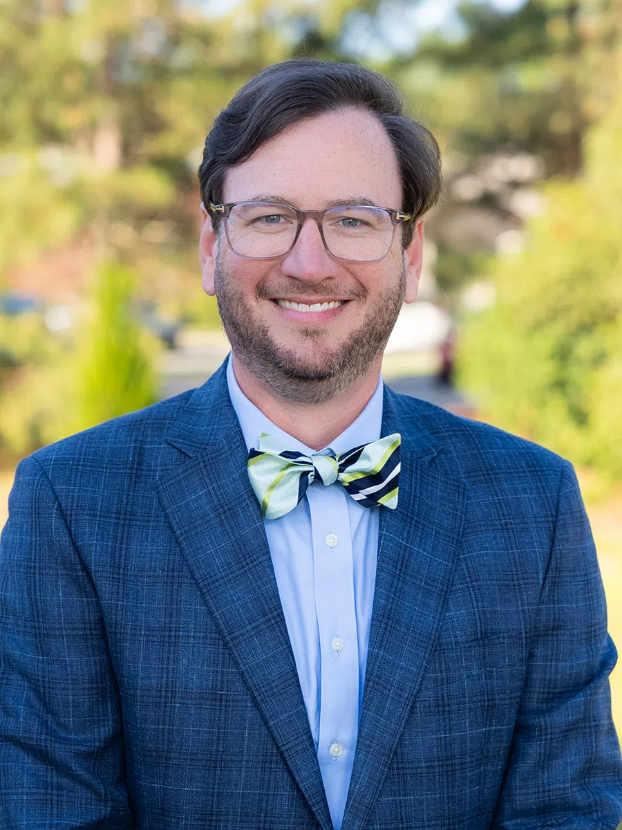 Smiling man with glasses wearing a blue checkered suit and striped bow tie outdoors with blurred greenery background