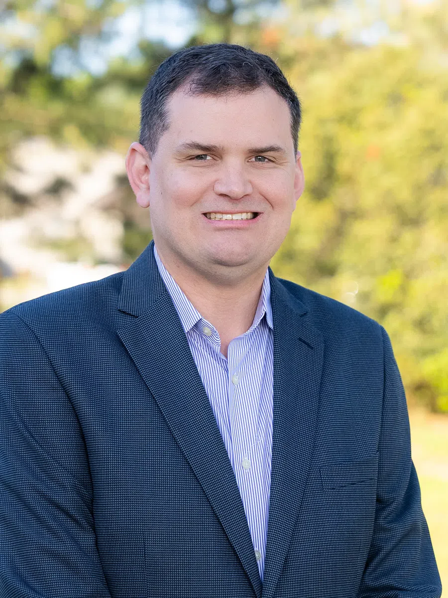 Smiling man in blue blazer and striped shirt posing outdoors with blurred green trees background