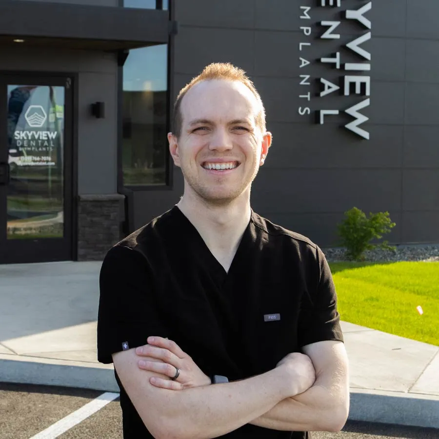 Smiling male dentist in black scrubs standing with arms crossed outside Skyview Dental clinic on a sunny day