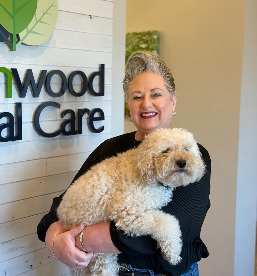 Smiling woman holding fluffy cream-colored dog at a veterinary clinic with sign in background.