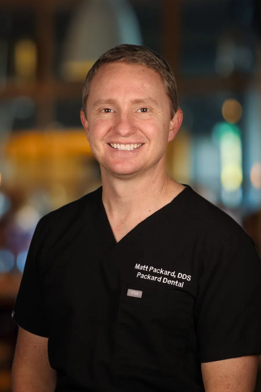 Smiling male dentist wearing black scrubs with Packard Dental logo in a softly lit background.