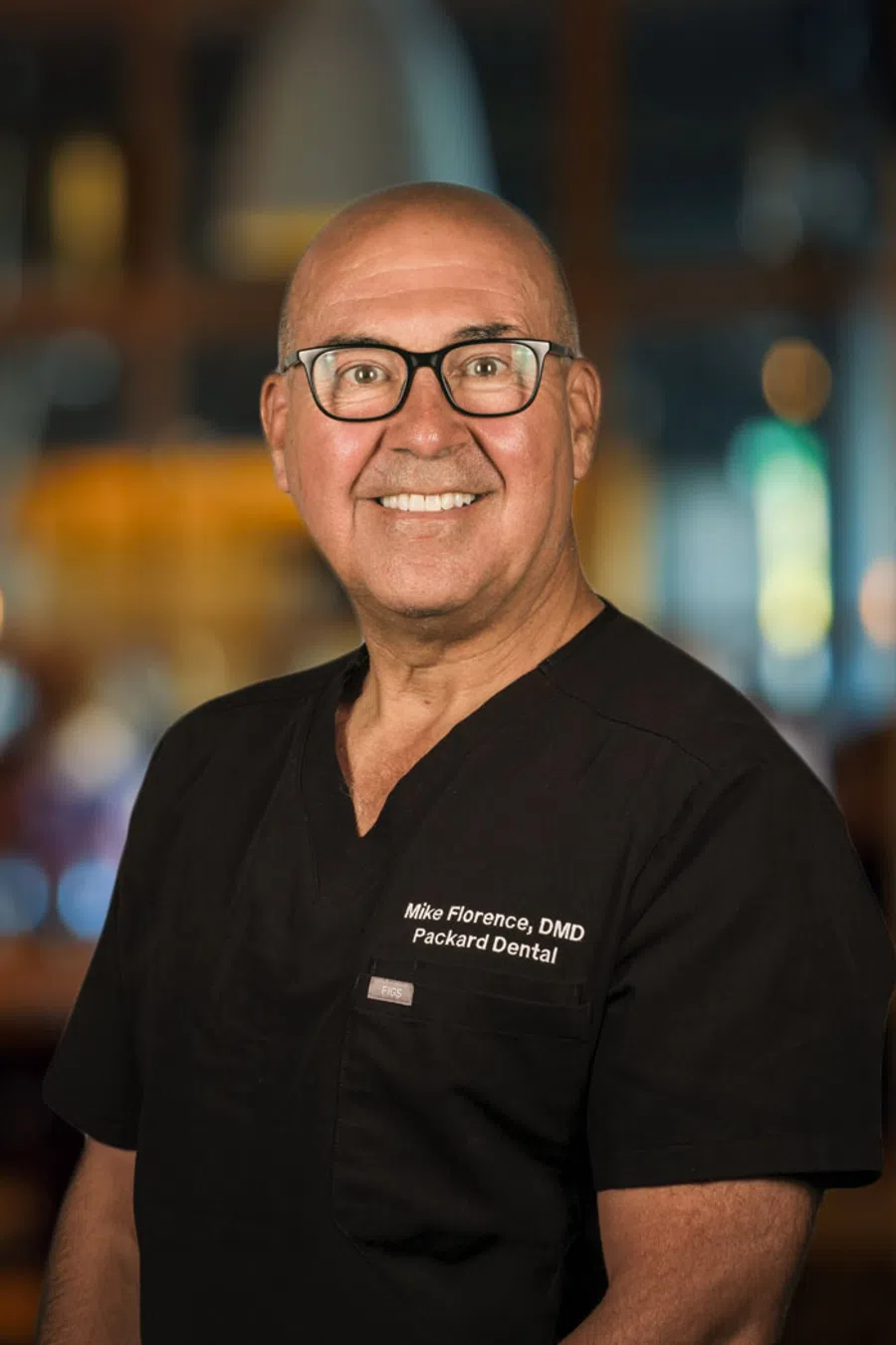 Smiling male dentist wearing black scrubs with glasses, posing against a blurred indoor background.