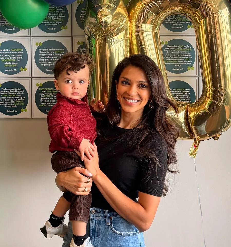 Smiling woman holding toddler with gold 10 balloon and positive reviews on wall behind them