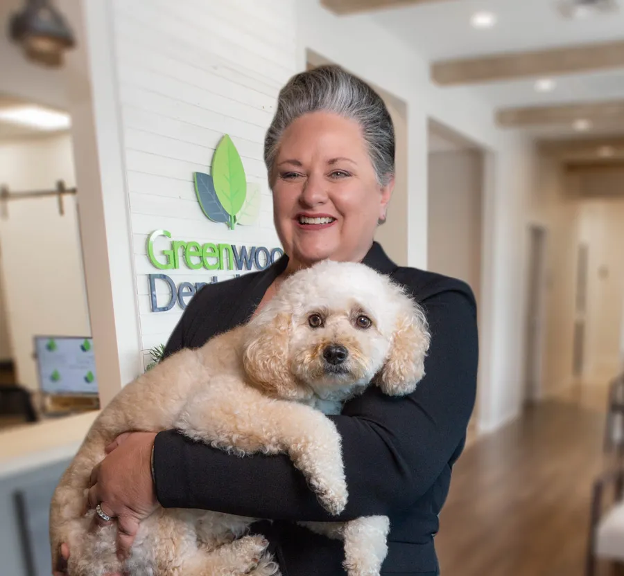 Woman with gray hair smiling and holding a fluffy white dog inside Greenwood Dental office.