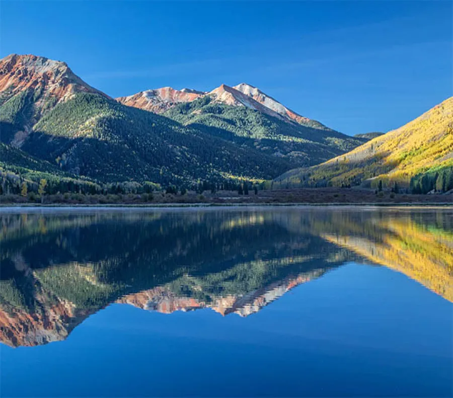 Mountain range and autumn trees reflected clearly in calm lake under blue sky