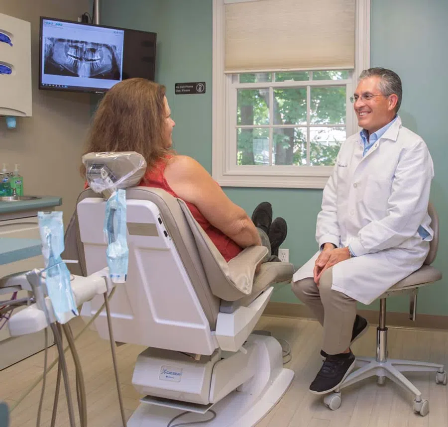 Dentist in white coat consulting with female patient in dental chair with tooth X-ray on screen.