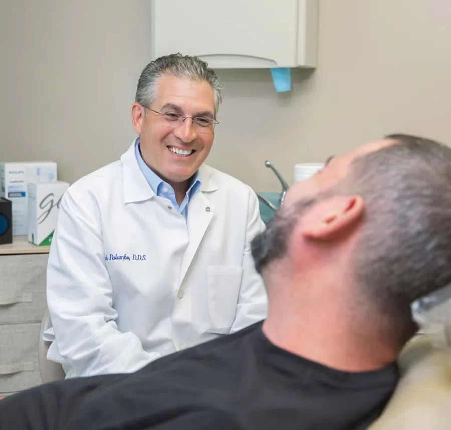 Smiling male dentist in white coat talks to male patient reclining in dental chair in clinic room.