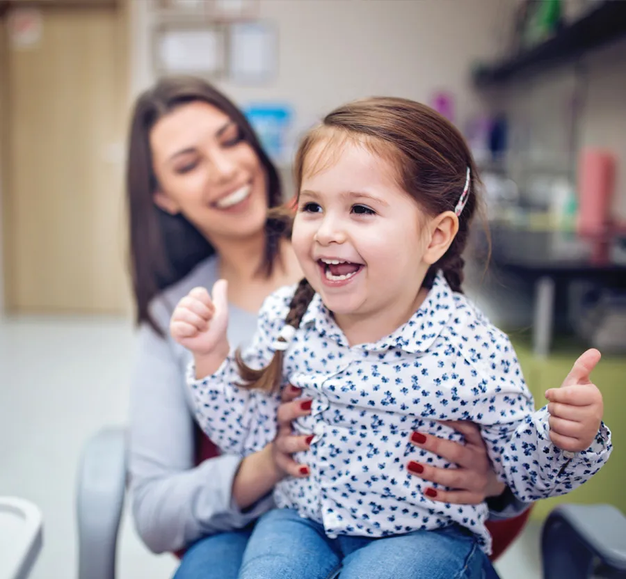 Happy young girl with braided hair sitting on woman's lap, both smiling and enjoying time together indoors.