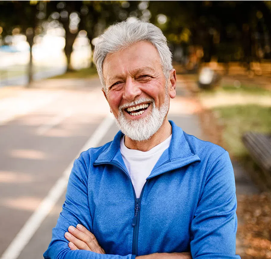 Smiling elderly man with white beard wearing a blue jacket standing outdoors on a path with blurred background