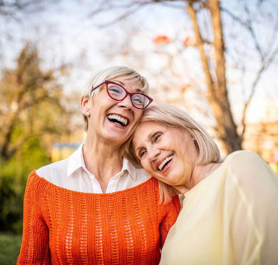 Two happy senior women outdoors, one with red glasses and orange sweater, sharing a joyful moment together.
