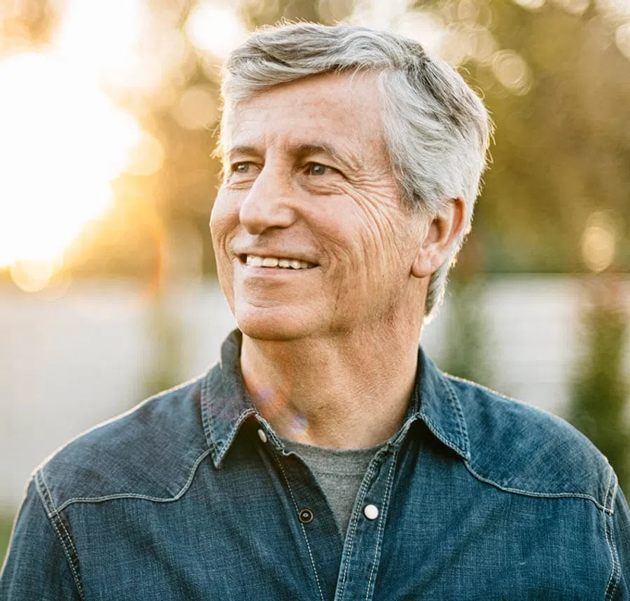 Smiling senior man with gray hair wearing a denim shirt standing outdoors in warm sunlight.