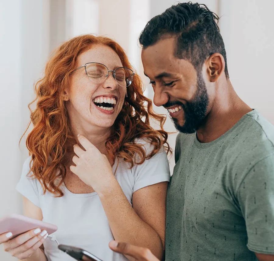 Happy young couple laughing while looking at their smartphones indoors in casual clothes.