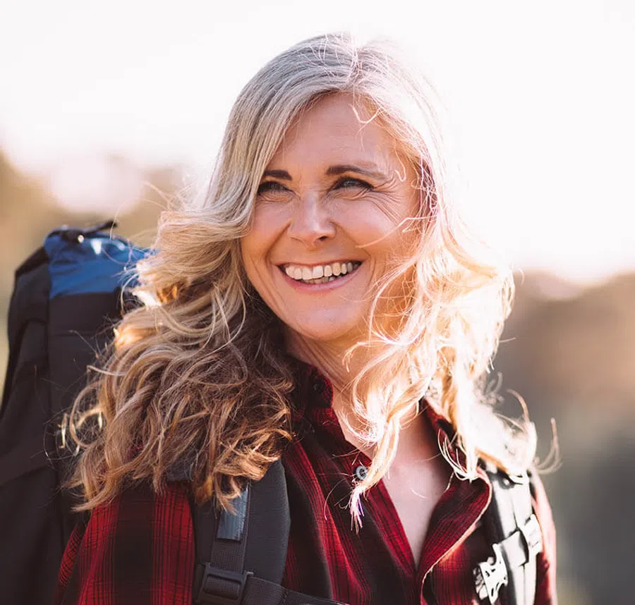 Smiling mature woman with long wavy hair wearing a red plaid shirt and backpack in bright outdoor setting