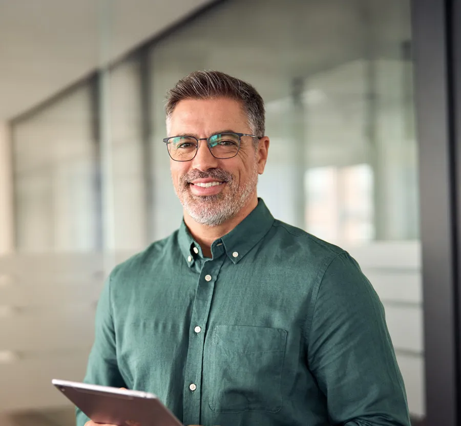 Smiling middle-aged man in glasses and green shirt holding tablet in modern office setting.