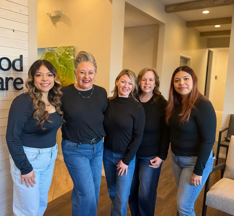 Group of five women standing indoors smiling, dressed in black tops and jeans, in a bright office setting.
