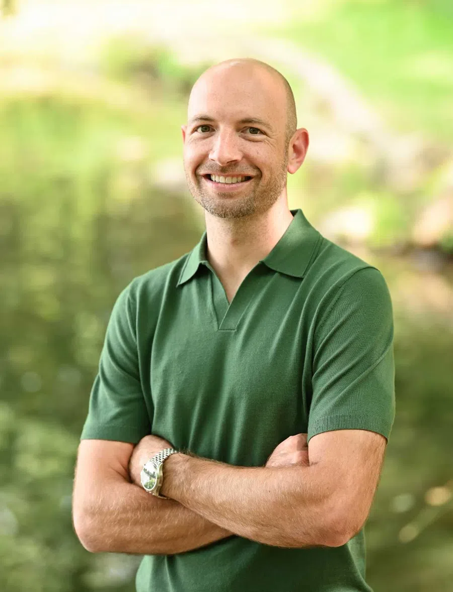 Smiling bald man in green polo shirt with arms crossed outdoors in a blurred natural green background.