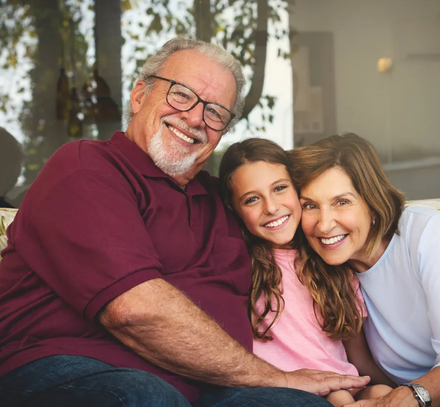 Smiling grandparents hugging their granddaughter seated between them on a couch in a cozy home setting