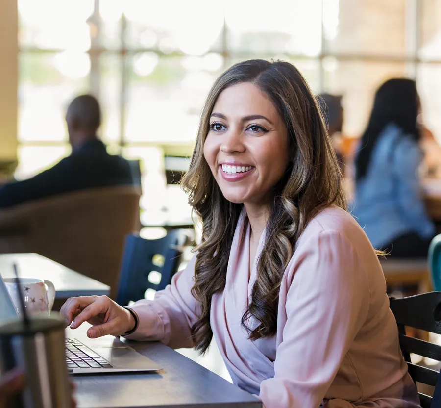 Smiling woman with long hair working on a laptop in a bright, busy cafe environment with other patrons.
