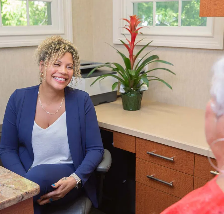 Smiling woman in blue blazer sitting and talking with another person in a bright office with plants and windows.