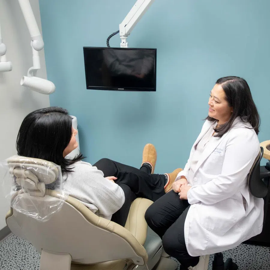 Dentist in white coat consulting with patient seated in dental chair in a modern clinic room.