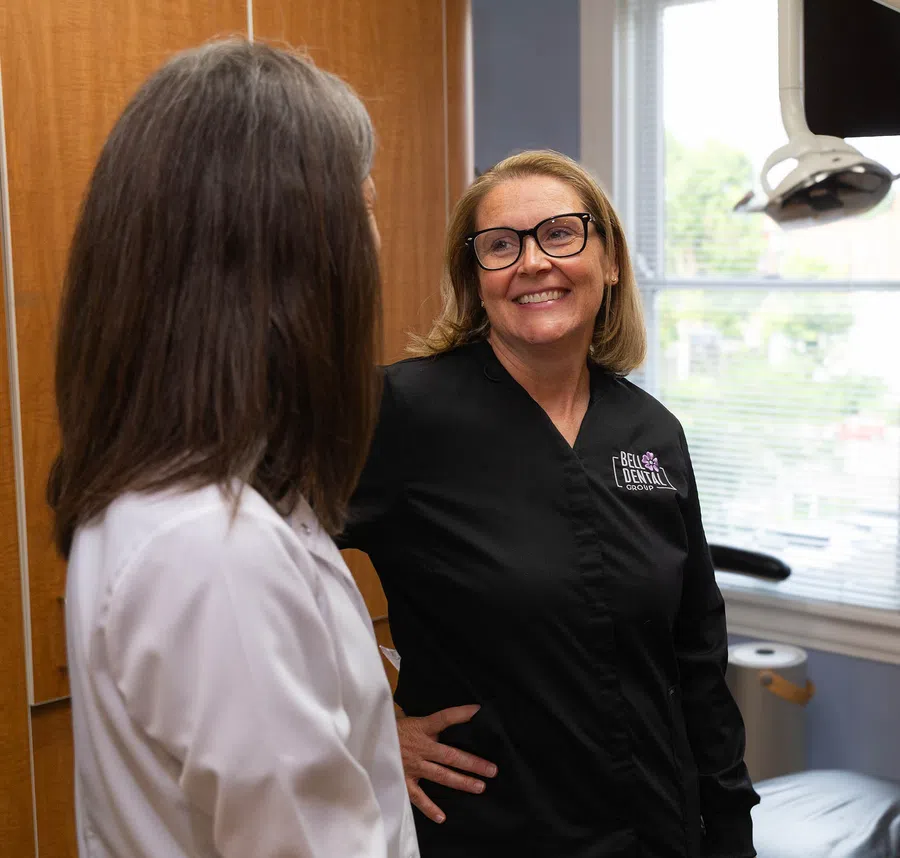 Two women in dental office uniforms talking in a bright room with dental equipment and window blinds.